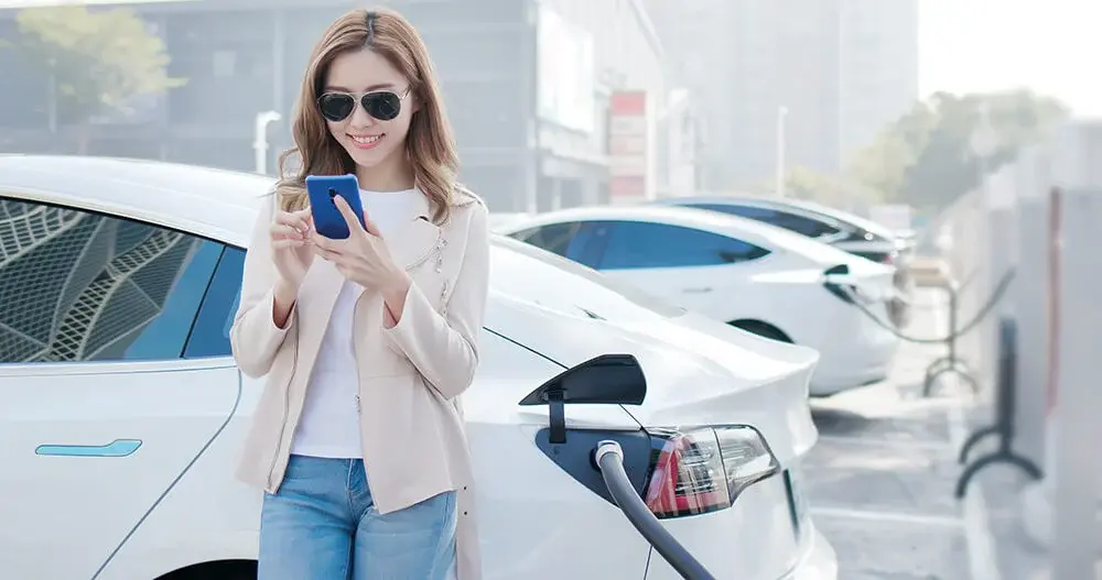 A person using a smartphone next to a white electric vehicle at a charging station.