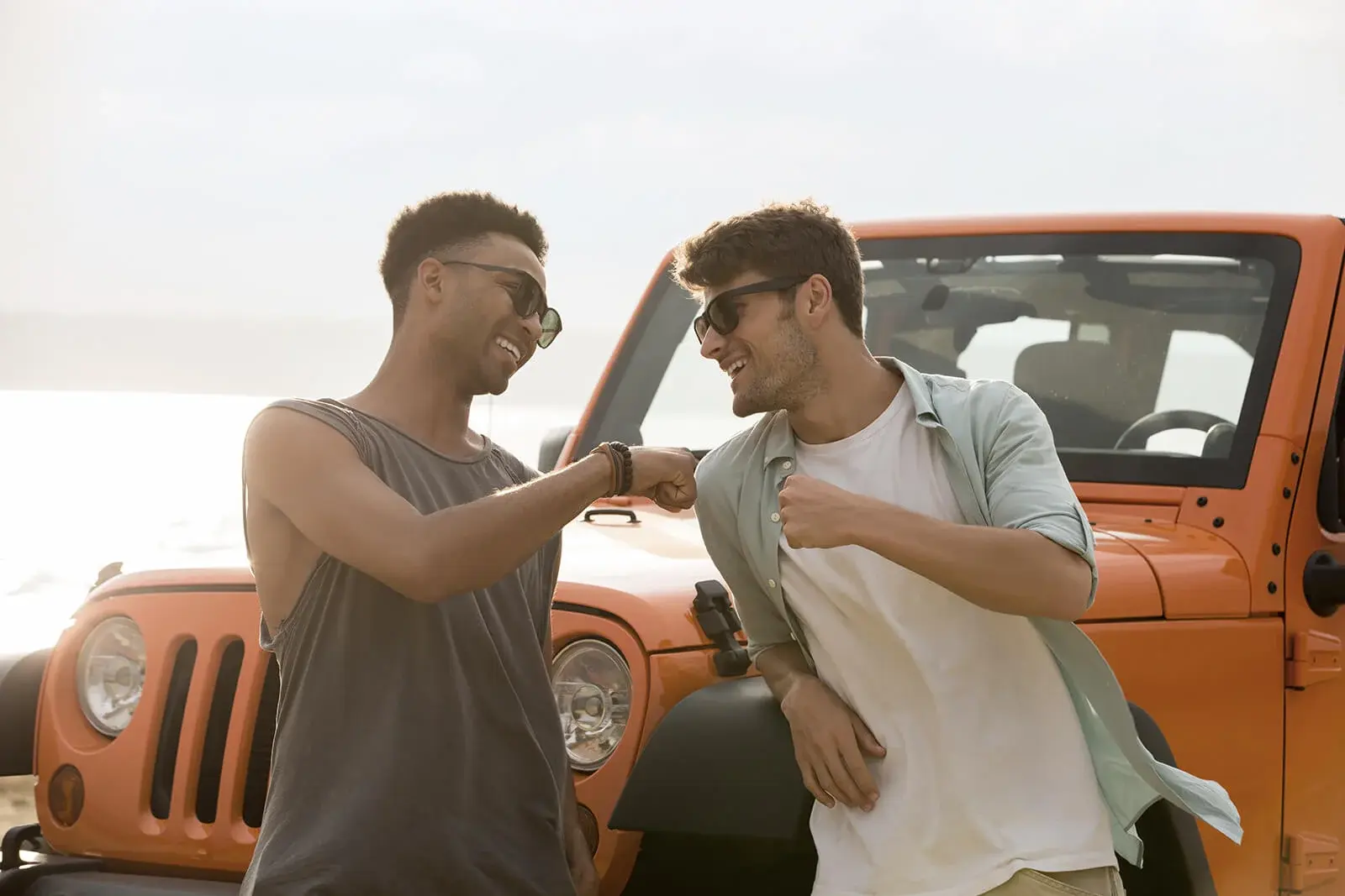 Two friends fist-bumping in front of an orange SUV on a bright day.